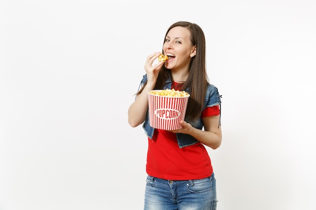 Retrato de joven sonriente hermosa mujer morena en ropa casual viendo una película, sosteniendo un cubo de palomitas de maíz, comiendo, disfrutando aislado sobre fondo blanco. Emociones en el concepto de cine.