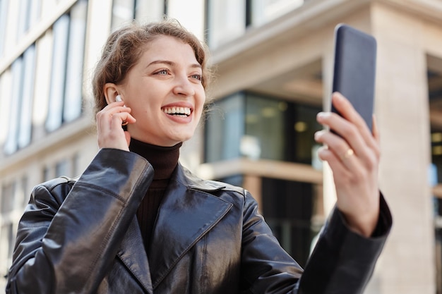 Retrato de una joven sonriente llamando por video chat en la ciudad y ajustando los auriculares inalámbricos