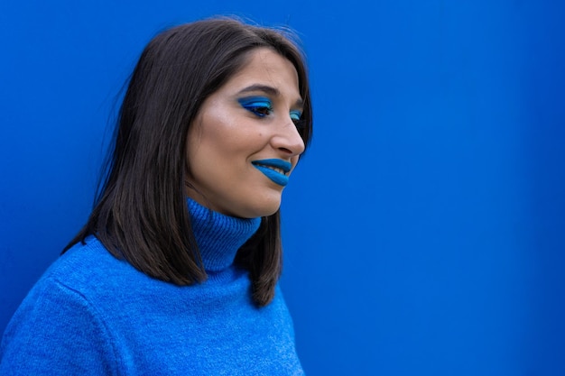 retrato de una joven sonriente con maquillaje de ojos azules y lápiz labial y suéter azul sobre un fondo azul.