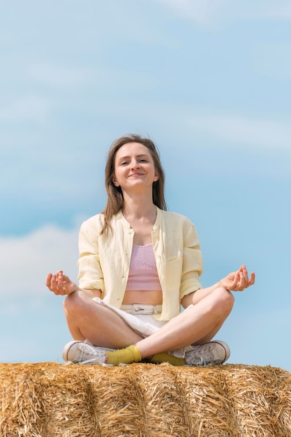 Retrato de una joven sonriente meditando sentada en el heno contra el cielo azul Unidad con la naturaleza