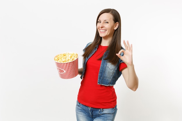 Retrato de joven sonriente mujer morena atractiva en ropa casual viendo una película, sosteniendo un cubo de palomitas de maíz y mostrando un signo bien aislado sobre fondo blanco. Emociones en el concepto de cine.