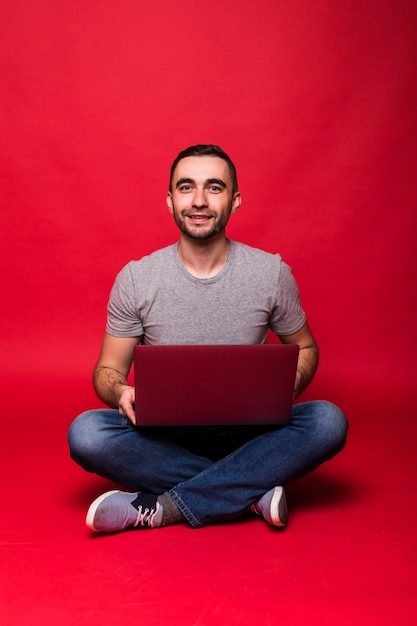 Retrato de un joven sonriente con ordenador portátil mientras está sentado en el suelo aislado sobre fondo rojo.