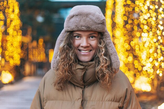 Retrato de una joven sonriente con el pelo rizado con sombrero de piel en la calle de invierno decorada con luces