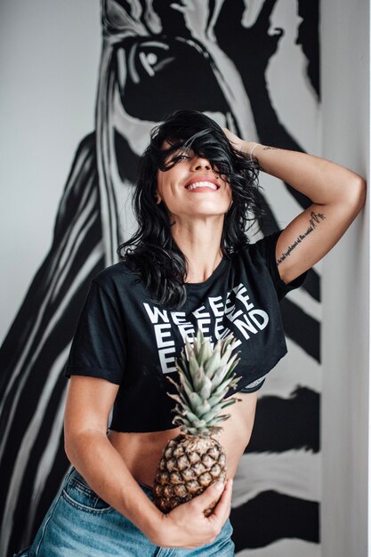 Foto retrato de una joven sonriente con una piña de pie contra la pared