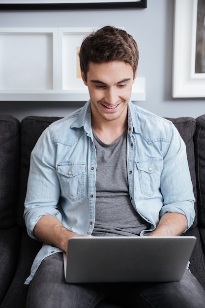 Retrato de un joven sonriente que trabaja en la computadora portátil mientras está sentado en el sofá en casa