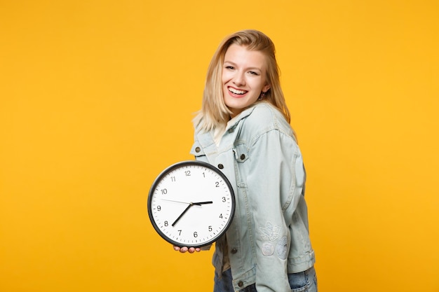 Retrato de una joven sonriente con ropa casual de denim mirando una cámara, sosteniendo un reloj redondo aislado en un fondo de pared naranja amarillo brillante en el estudio. Concepto de estilo de vida de las personas. Simulacros de espacio de copia.