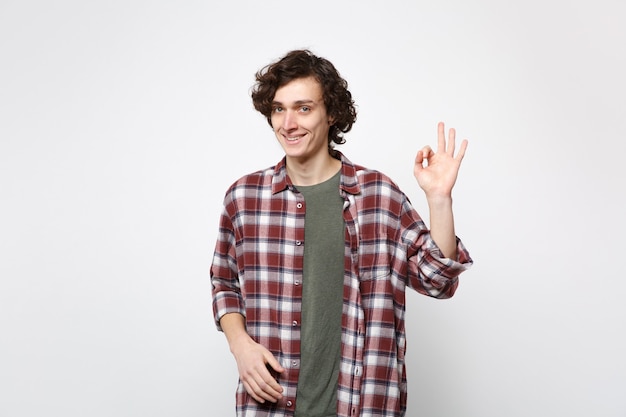 Retrato de joven sonriente en ropa casual de pie, mirando a cámara mostrando gesto OK aislado sobre fondo de pared blanca en estudio. Personas sinceras emociones, concepto de estilo de vida. Simulacros de espacio de copia.