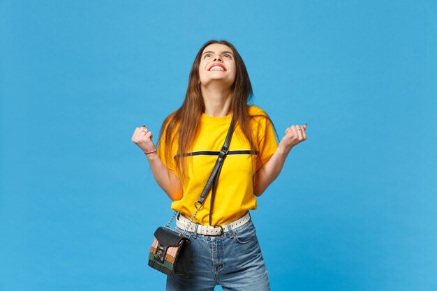 Retrato de una joven sonriente con ropa casual vívida con una bolsa de cuerpo cruzado apretando los puños como un ganador mirando hacia arriba aislado en un fondo azul brillante. Concepto de estilo de vida de las personas. Simulacros de espacio de copia.