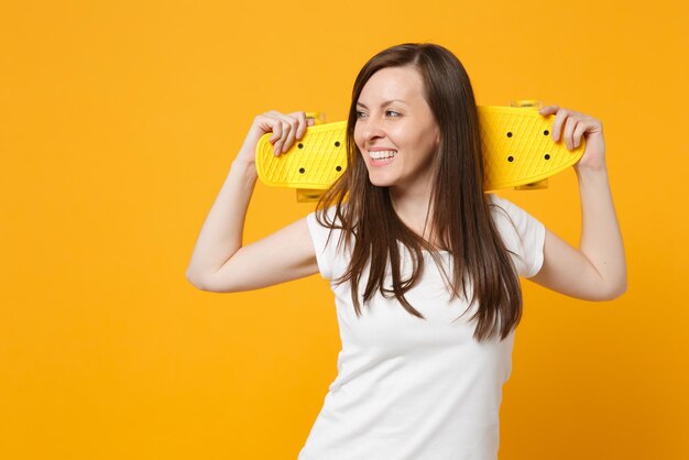 Retrato de una joven sonriente con ropa informal blanca mirando a un lado, sosteniendo una patineta amarilla aislada en un fondo naranja amarillo brillante en el estudio. Concepto de estilo de vida de las personas. Simulacros de espacio de copia.