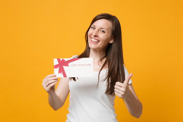 Retrato de una joven sonriente con ropa informal blanca que muestra el pulgar hacia arriba, sosteniendo un certificado de regalo aislado en un fondo naranja amarillo brillante en el estudio. Concepto de estilo de vida de las personas. Simulacros de espacio de copia.