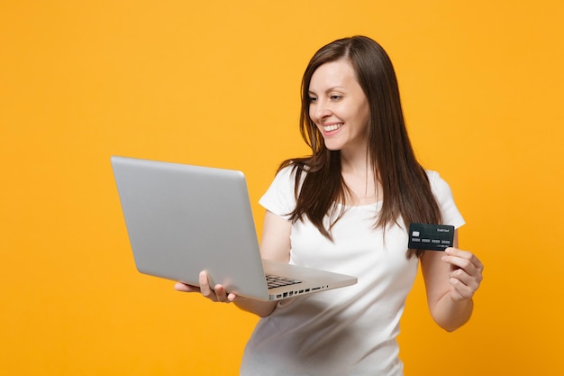 Retrato de una joven sonriente con ropa informal blanca sosteniendo un ordenador portátil, tarjeta bancaria de crédito aislada en un fondo de pared naranja amarillo en el estudio. Concepto de estilo de vida de las personas. Simulacros de espacio de copia.