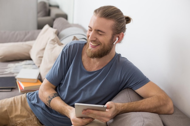 Retrato de un joven sonriente sentado en un sofá gris con auriculares y la tableta en las manos en casa