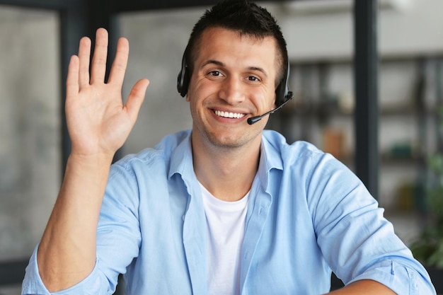 Retrato de un joven sonriente servicios de apoyo para empleados en el auricular mirando a la cámara muestra un gesto de saludo, comunicación en línea, consulta, videollamada, reunión