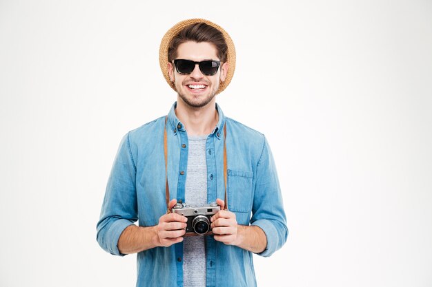 Retrato de joven sonriente con sombrero y gafas de sol con cámara de fotos de época antigua sobre fondo blanco.