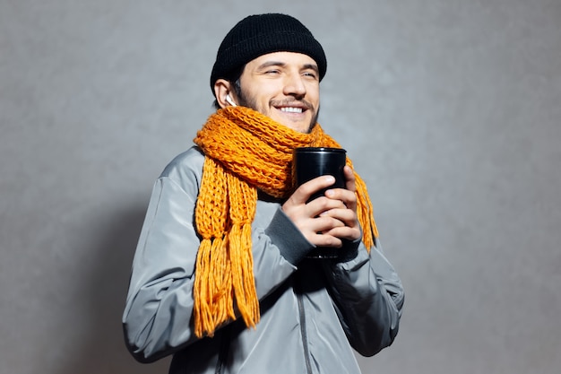 Retrato de joven sonriente con taza termo de café en las manos, con auriculares inalámbricos