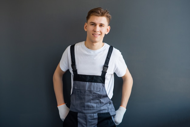 Retrato de un joven, sonriente, trabajador profesional en overoles grises y camiseta blanca, de pie sobre un fondo gris.