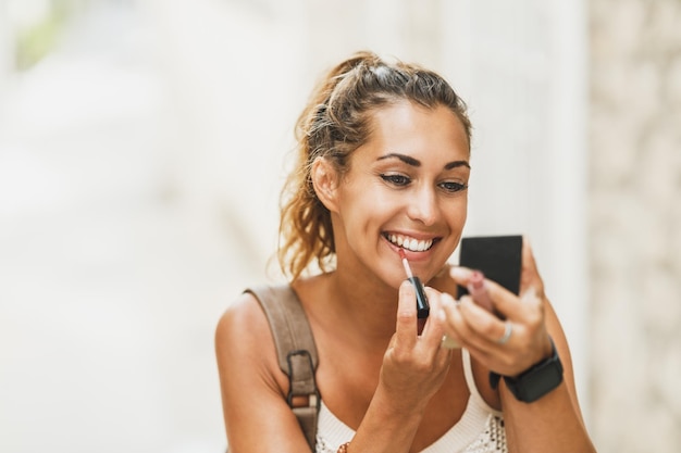 Retrato de una joven sonriente usando espejo de mano y aplicando lápiz labial mientras disfruta de unas vacaciones de verano.