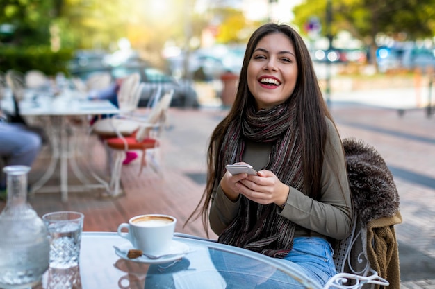 Retrato de una joven sonriente usando un teléfono móvil mientras está sentada en una cafetería en la acera