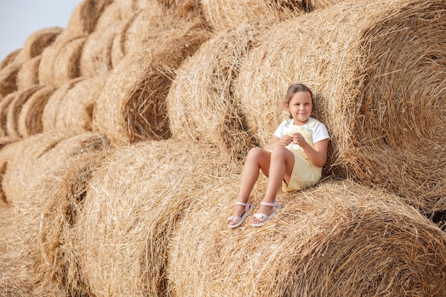 Retrato de una joven sonriente con vestido de verano sentada en un pajar alto y apoyándose en otro y muchos otros pajares en el fondo Divirtiéndose lejos de la ciudad en un campo lleno de heno dorado