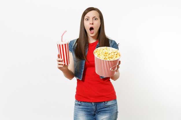 Retrato de joven sorprendida preocupada en ropa casual viendo una película, sosteniendo un cubo de palomitas de maíz y un vaso de plástico de refresco o cola aislado sobre fondo blanco. Emociones en el concepto de cine.