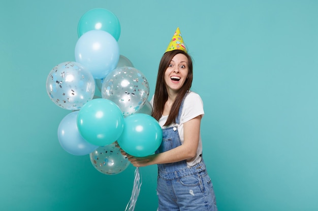Retrato de una joven sorprendida con ropa de denim, sombrero de cumpleaños celebrando, sosteniendo coloridos globos de aire aislados en un fondo azul turquesa de la pared. Fiesta de cumpleaños, concepto de emociones de la gente.