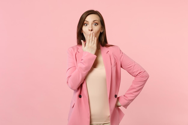 Retrato de una joven sorprendida sorprendida con una chaqueta de pie, cubriendo la boca con la palma aislada en un fondo de pared rosa pastel en el estudio. Emociones sinceras de la gente, concepto de estilo de vida. Simulacros de espacio de copia.