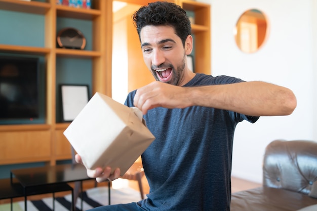 Retrato de un joven sorprendido abriendo una caja de regalo mientras está sentado en el sofá en casa.