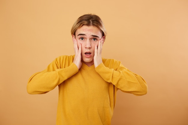 Retrato de joven sorprendido rubio vestido con suéter amarillo está planteando en el estudio sobre fondo beige.