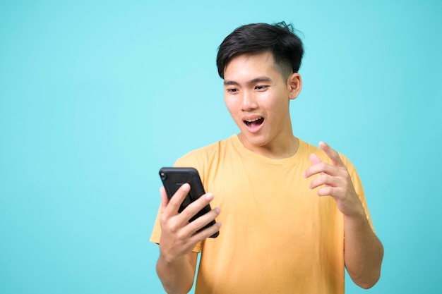 Retrato de un joven sorprendido mediante teléfono móvil aislado sobre fondo azul.