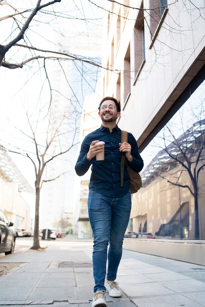 Retrato de joven sosteniendo una taza de café mientras camina al aire libre en la calle. Concepto urbano y de estilo de vida.