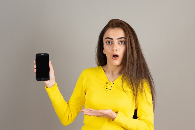 Retrato de joven sosteniendo el teléfono celular con expresión de sorpresa en la pared gris.