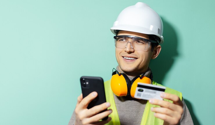 Foto retrato de joven trabajador ingeniero de la construcción con equipo de seguridad, usando un teléfono inteligente y una tarjeta bancaria de crédito sobre un fondo de color aqua menthe.