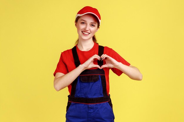 Retrato de una joven trabajadora de aspecto agradable parada y mostrando un gesto de corazón, expresando felicidad, usando overoles y gorra roja. Disparo de estudio interior aislado sobre fondo amarillo.