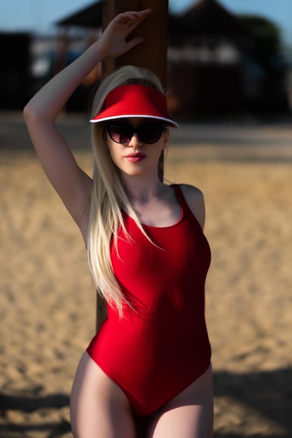 Retrato de una joven con un traje de baño rojo de una pieza, gafas de sol y una gorra roja posando en la playa apoyada en un soporte de madera Concepto de vacaciones vacaciones de verano