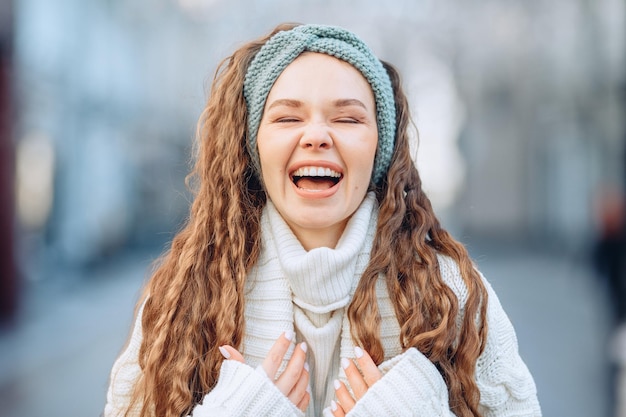 Retrato de una joven con un traje de punto blanco y una banda azul. Fondo borroso de la ciudad. Tonterías en cámara. Ojos cerrados, cerrados con fuerza y sonrisa brillante. El concepto de emociones positivas.