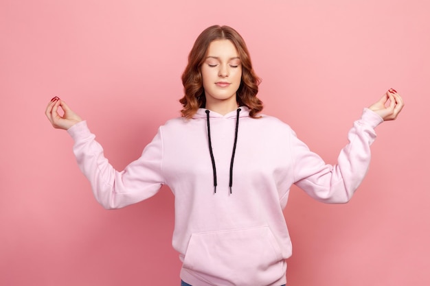 Retrato de una joven tranquila con cabello castaño encapuchado de pie con los ojos cerrados, meditando con gesto de mudra, mente pacífica. Disparo de estudio interior aislado sobre fondo rosa