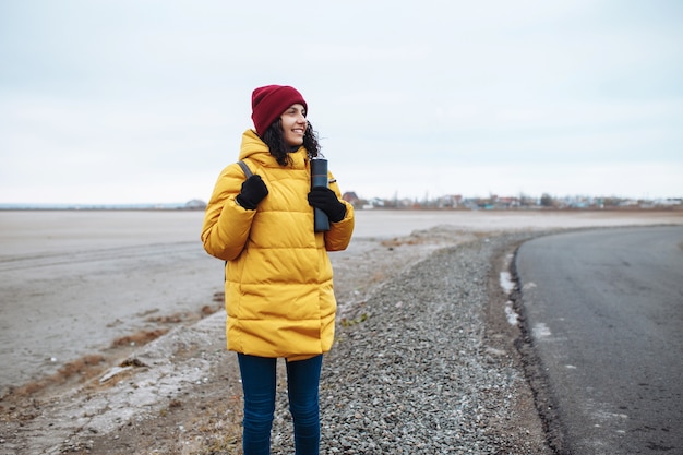 Retrato de una joven turista con una mochila caminando por la calle lateral entre las vastas tierras bajas del valle de invierno vacío. Viajero mujer vestida con chaqueta amarilla y sombrero rojo. Autostop, concepto de viaje.