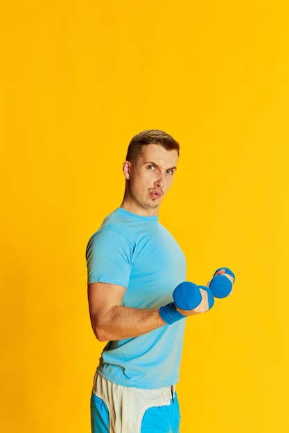 Retrato de un joven con uniforme azul entrenando con pesas posando aislado sobre un fondo amarillo Concepto de deporte y emociones