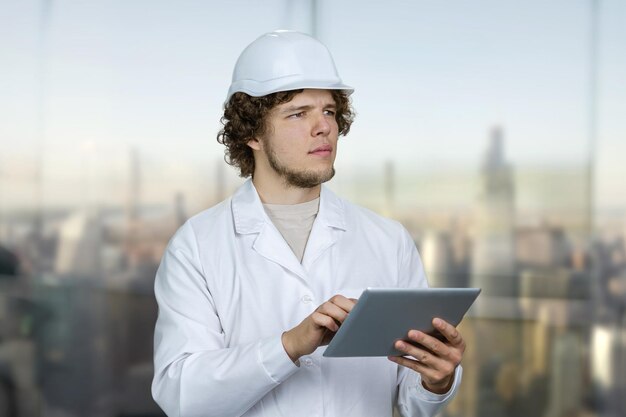 Retrato de un joven en uniforme blanco y casco blanco sosteniendo una tableta dispositivo de PC borroso paisaje urbano