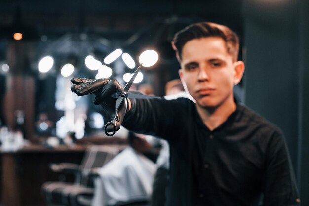 Retrato de un joven vestido de negro que está parado adentro en una barbería y sosteniendo unas tijeras