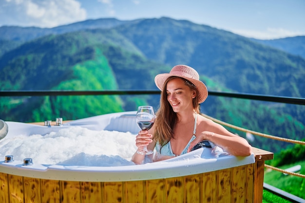 Retrato de joven viajero despreocupado feliz sonriente niña relajándose con una copa de vino en el tubo caliente durante el disfrute de la vida de vacaciones feliz momento de viaje en el contexto de grandes montañas verdes