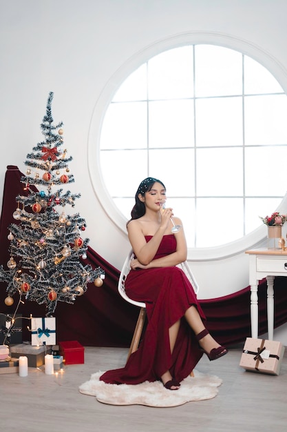 Retrato de una jovencita acogedora sentada y bebiendo vino vestida de rojo en una sala de estar navideña decorada en el interior