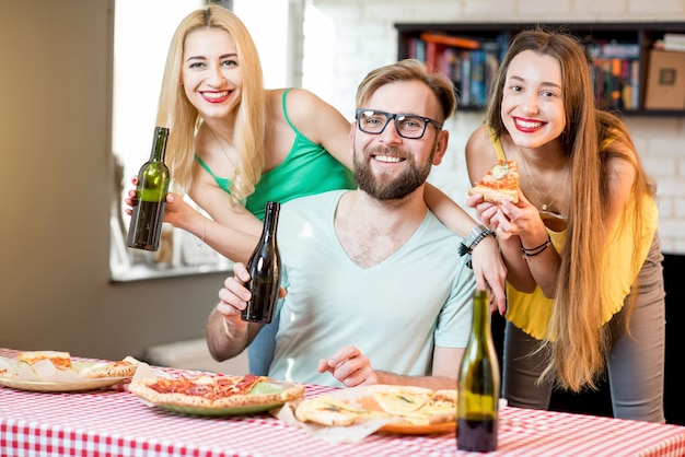 Retrato de los jóvenes amigos vestidos casualmente con coloridas camisetas almorzando con pizza y cerveza en casa