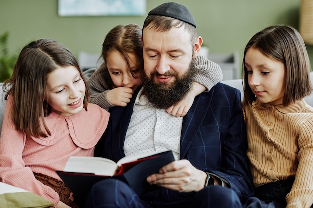 Foto retrato de un judío ortodoxo que usa kipá mientras lee un libro a tres niños