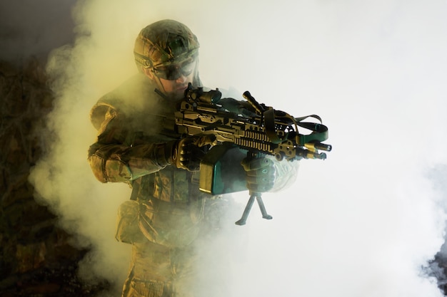 Retrato de jugador de airsoft en equipo profesional con ametralladora en edificio abandonado en ruinas. Soldado con armas en guerra en humo y niebla.