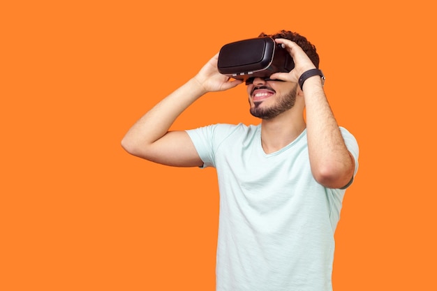 Retrato de jugador feliz, hombre moreno con barba en camiseta con auriculares vr, jugando al juego de realidad virtual con una sonrisa en la cara, tecnología innovadora. tiro del estudio aislado sobre fondo naranja