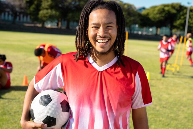 Retrato de un jugador masculino birracial con rastas sosteniendo una pelota de fútbol y riéndose en el patio de juegos