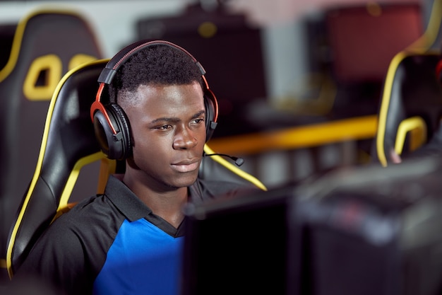 Retrato de un jugador masculino de cybersport africano enfocado con audífonos jugando videojuegos, participando en un torneo de eSport mientras está sentado en un club de juegos o cibercafé