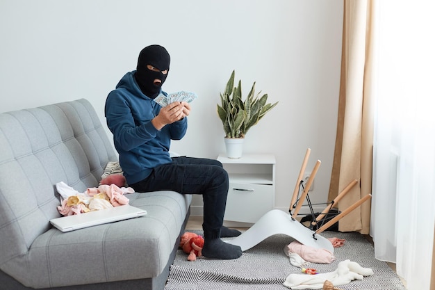 Retrato de ladrón sorprendido sorprendido hombre vestido con sudadera con capucha azul y pasamontañas negro sentado en el sofá en casa, sorprendido al encontrar una gran suma de dinero, expresando asombro.