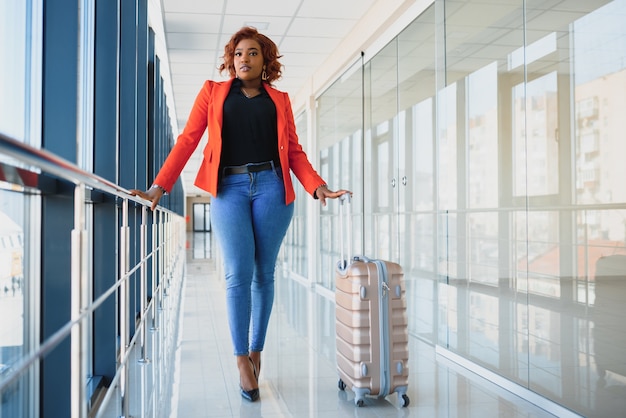 Retrato lateral de corpo inteiro de jovem negra caminhando com uma mala no aeroporto